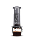 Aeropress coffee maker with a dark gray body and clear chamber, shown with brewed coffee dripping into a clear glass cup on a white background.