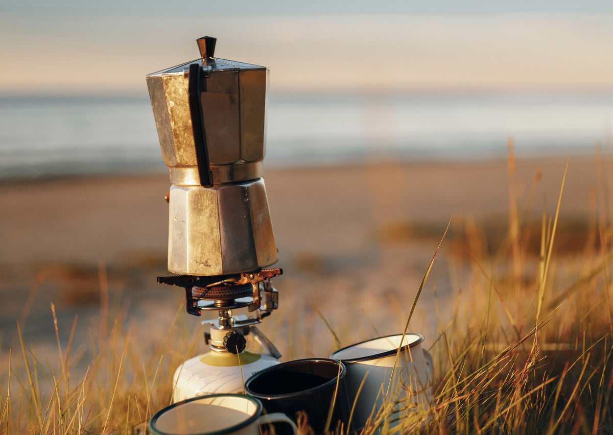 Moka pot with cups on a camping stove by a body of water
