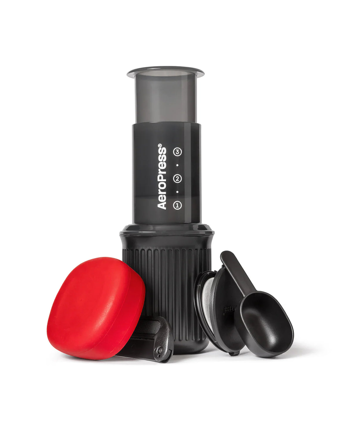 A portable AeroPress coffee maker in black with a translucent gray top chamber, shown with a bright red lid and two black measuring scoops on a clean white background.