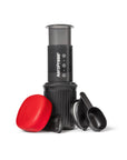 A portable AeroPress coffee maker in black with a translucent gray top chamber, shown with a bright red lid and two black measuring scoops on a clean white background.