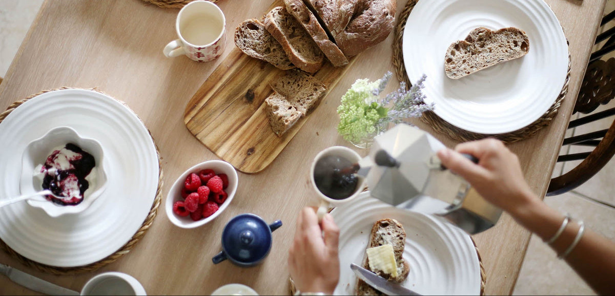 Overhead view of a breakfast table setting with sliced bread on a wooden cutting board, white plates, bowls of jam and butter, cups, and two visible hands serving food at a light wood dining table.