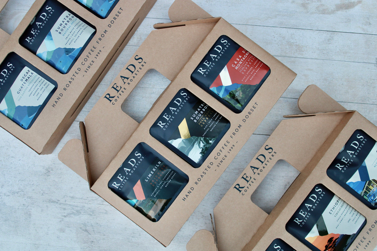 Several brown kraft paper coffee gift boxes with cutout handles are arranged on a light gray wood surface. Each box contains multiple small coffee cards showing different flavor and origin labels, and the brand name READS is clearly printed on the boxes.