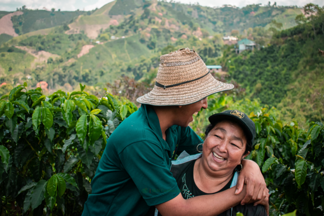 Two smiling women embracing on a lush green hillside, with one wearing a straw hat and teal shirt and the other wearing a black cap. Rolling green hills fill the background.