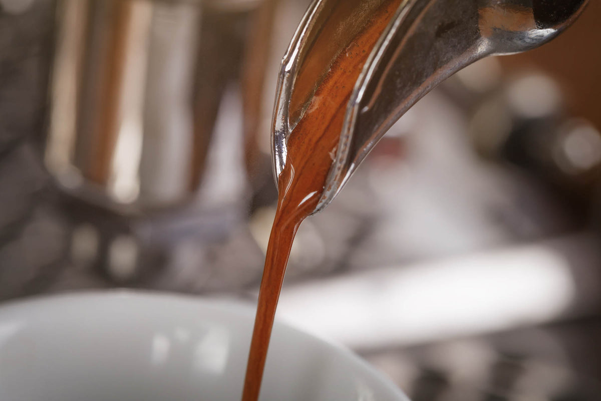 A close-up shot of espresso being poured from a metal portafilter into a white cup, with a blurred coffee maker in the background.