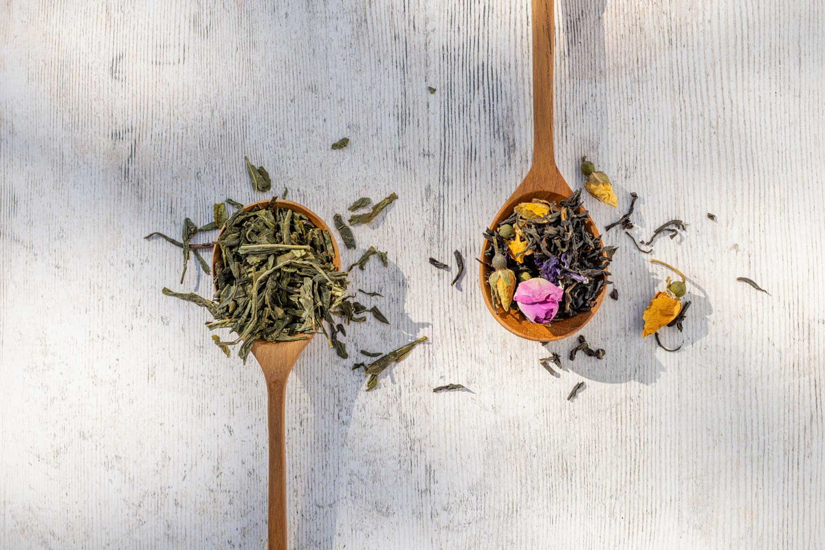 Two wooden spoons filled with loose-leaf tea on a light gray wooden surface. One spoon contains green tea leaves, and the other contains a multicolor herbal tea blend with dried flowers.