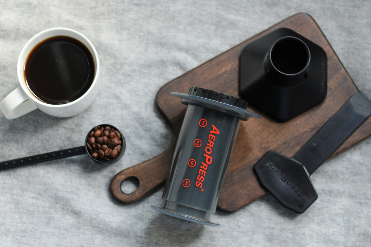 AeroPress coffee maker on a wooden board with a cup of coffee and coffee beans on a gray surface.
