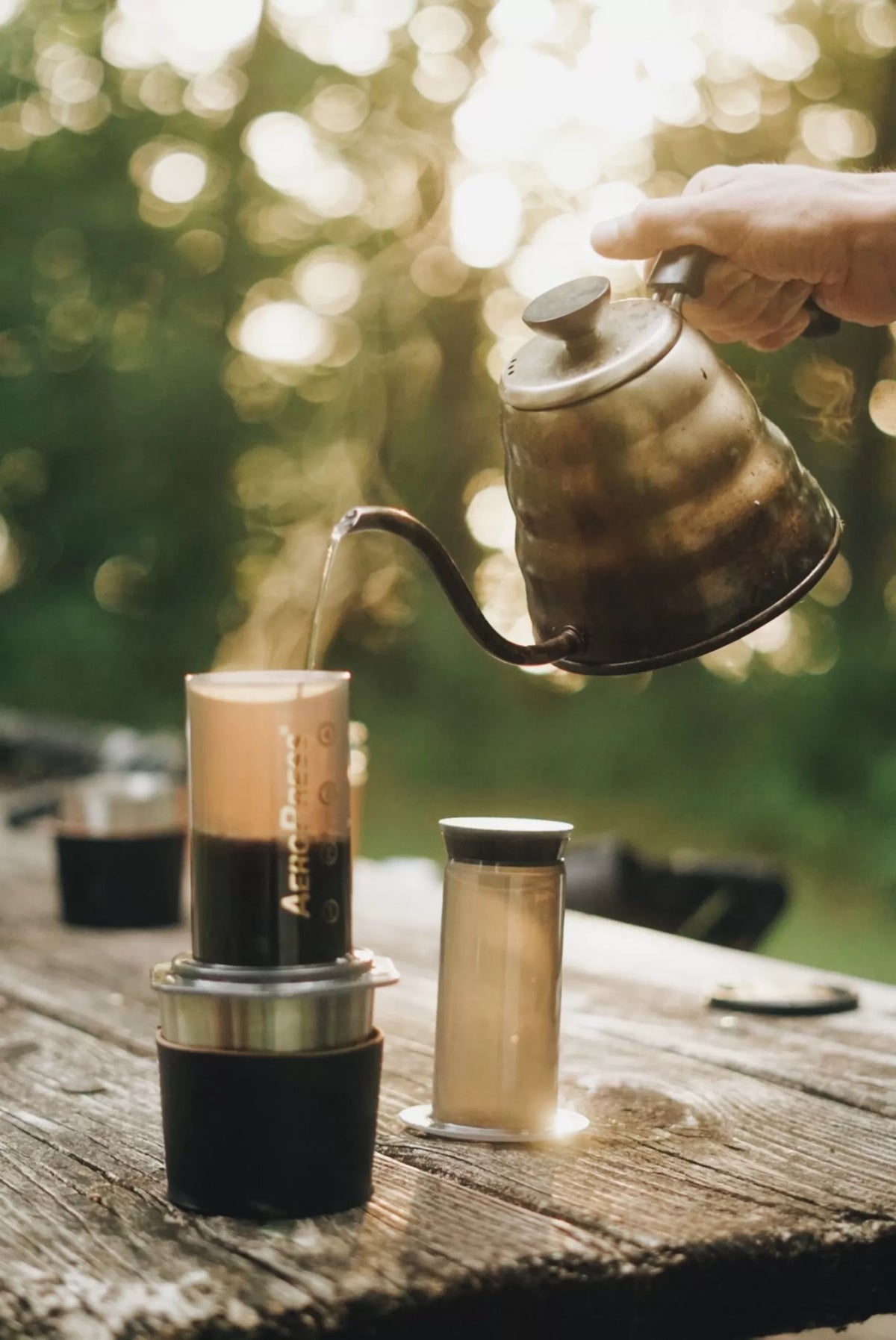 Person pouring coffee from a kettle into a portable coffee maker on a wooden surface with a blurred natural background.