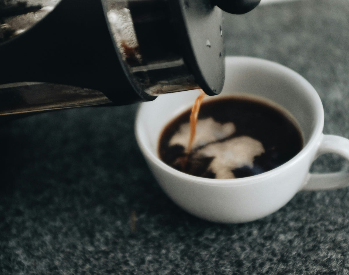 Coffee being poured from a French press into a white mug on a dark surface.