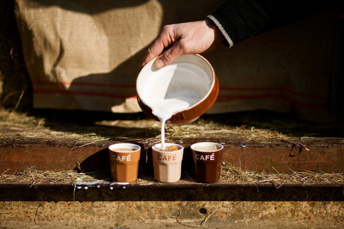 A hand pours white milk from a brown ceramic pitcher into three small ceramic cups labeled “CAFÉ” arranged on a rustic wooden surface outdoors.