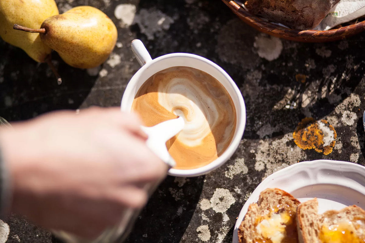 Cup of coffee with a latte art heart, surrounded by bread and pears on a rustic surface.