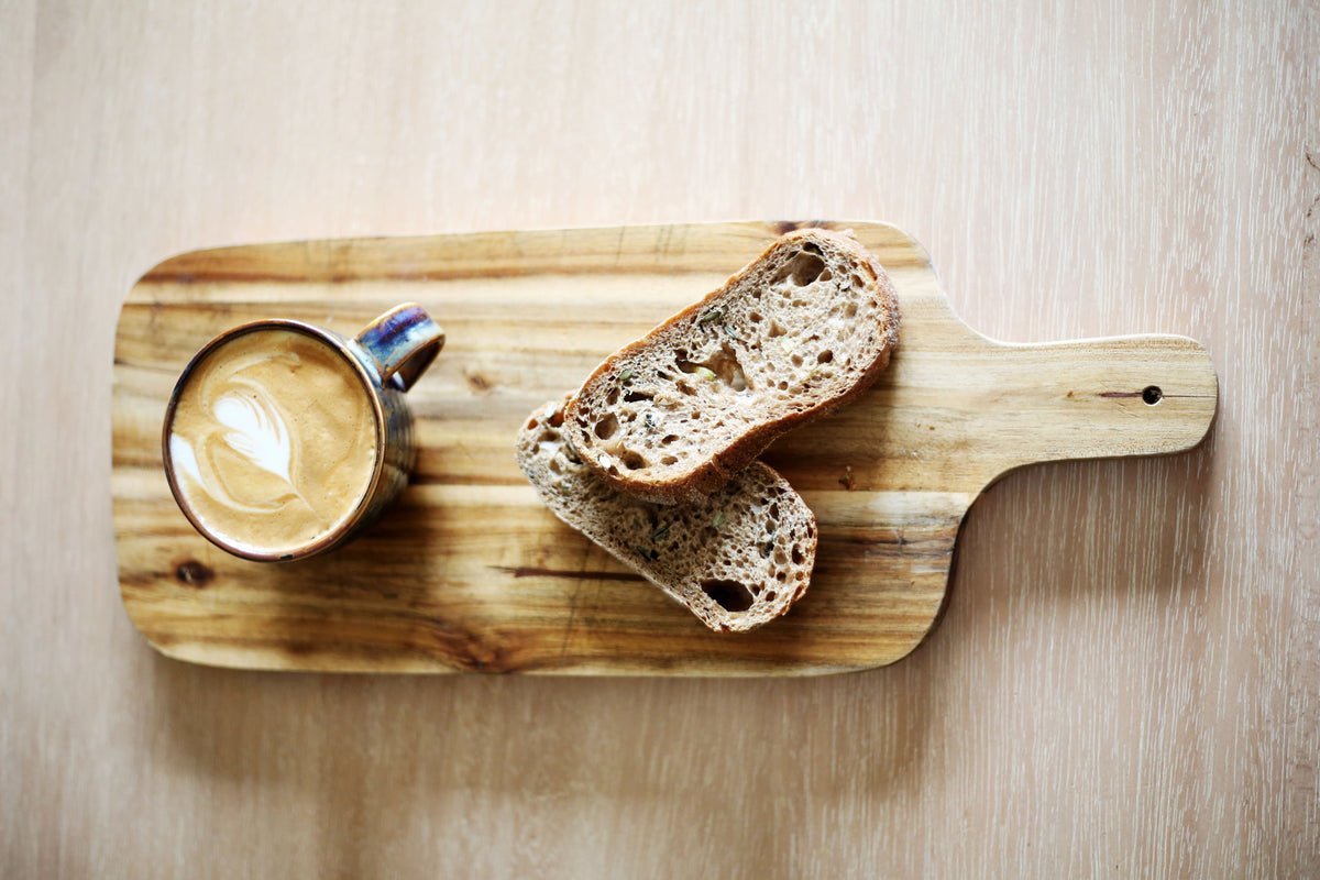 A top-down view of a rustic wooden cutting board on a light wood table, holding a small ceramic cup of latte with leaf art and two slices of artisanal sourdough bread.